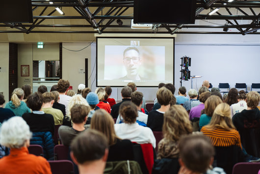 Projektion des per Videokonferenz zugeschalteten  Salzburger Vizebürgermeister Kay-Michael Dankl, davor Publikum von hinten 