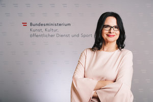 Staatssekretärin Andrea Mayer vor dem Logo des BMKÖS