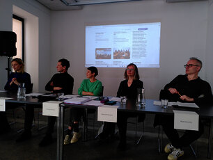 Zu sehen sind 5 Personen hinter einem Podiumstisch sitzen. Dahinter ist ein Text mit BIldern an die Wand projiziert, von dem am Foto aber nichts erkennbar oder lesbar ist.