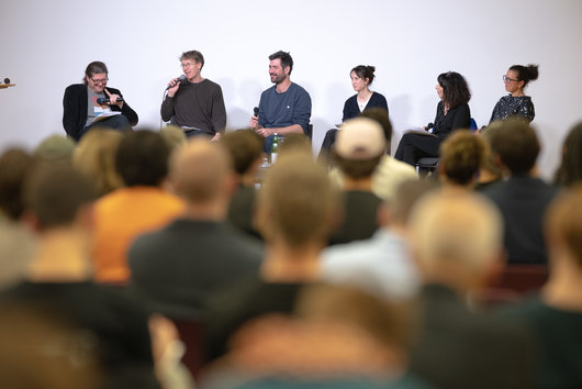 Die Diskussionsteilnehmer*innen am Podium sitzend, davor (von hinten fotografiert) Teile des Publiums