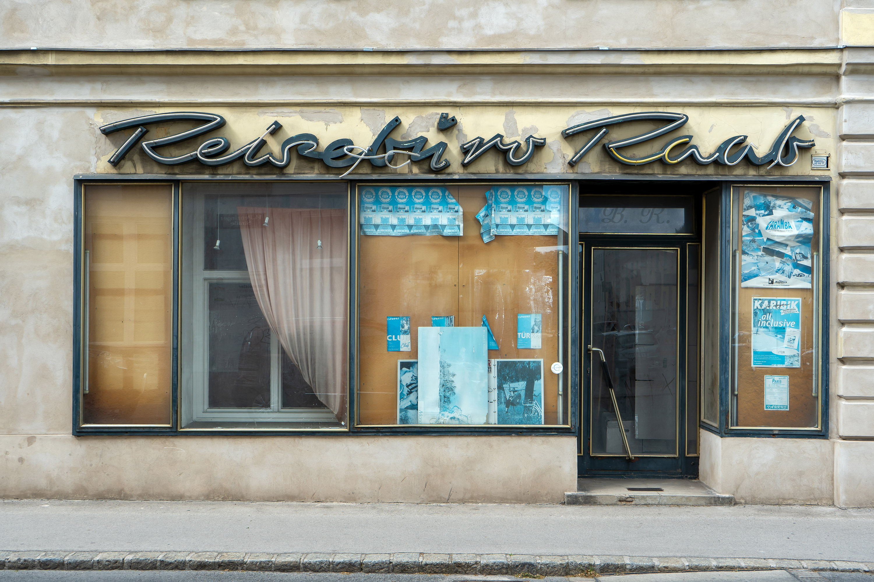 Foto eines leerstehenden Reisebüros, Firmenname "Reisebüro Raab" ist schon teilweise runtergefallen, Schaufenster sind großteils leer, ein paar ausgebleichte alte Plakate von Reiseveranstaltungen hängen noch. 